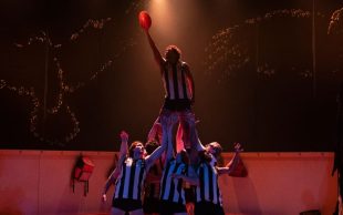 37. A group of men in AFL strips stand on stage and hold one of their group high above them. He has his arm upstretched holding an Australian Rules Football ball.