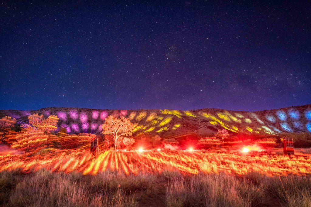 Light show projected onto the MacDonnell Ranges at Parrtjima 2023. Photo: Supplied. Orange, pink, yellow and blue light projects of First Nations patterns on the hilly surface of the MacDonnell Ranges under a starry sky. 