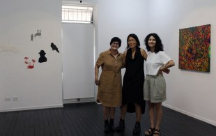 Three female artists stand in front of their artwork in the Woolloongabba Art Gallery.