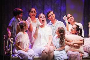 The Sound of Music. Image is a woman in a white nightgown sitting on a bed singing to the seven children sitting and standing around her.