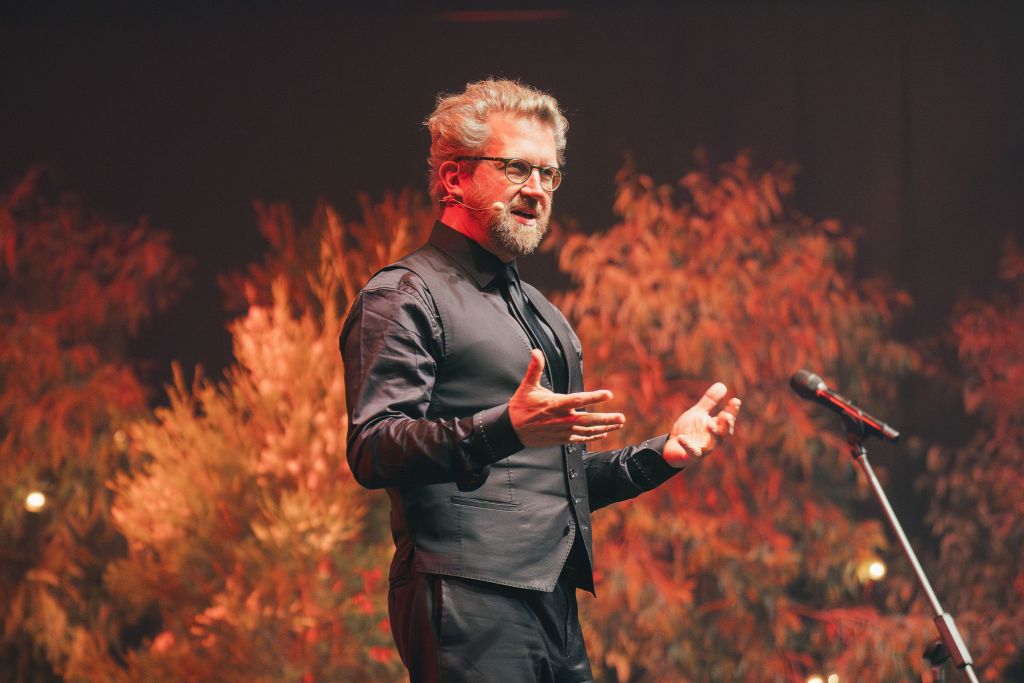 Man on stage in a black shirt, trousers and waistcoat, speaking to microphone with brown red bush behind him. Iain Grandage.