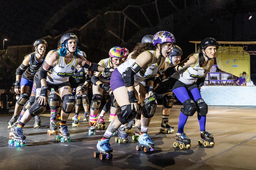 Roller Coaster. A group of roller derby women are skating towards the right of the frame, bending over. They are wearing helmets and other protective gear.