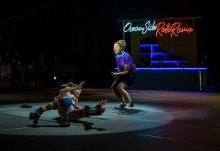 Roller Coaster. A dark stage has a backdrop of neon lights in the top right hand corner of the frame, saying 'Ocean Side Rolla Rama'. In a spotlight in the centre of the stage are two women, one wearing skates and multi-coloured socks has crashed to the ground. Behind her a commentator in a purple shirt with a headset mic is crouching and shouting - is she cheering or jeering? We don't know.
