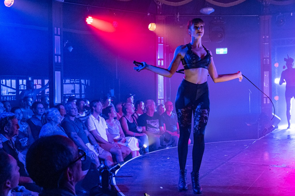 Rouge. A woman in black lacy tights and leather struts across a red spotlit stage holding a whip behind her.
