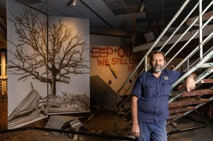 Cyclone Tracy - 50 Years On. A bearded man in a blue shirt and trousers, leans on a stair handrail in a gallery space. Behind him are flats with images on, most clearly a large tree surrounded by debris of corrugated iron following a cyclone.
