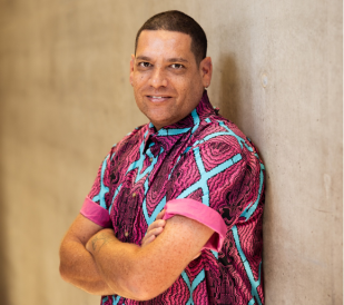 Fondation Cartièr. A man of Indigenous appearance is wearing a brightly coloured purple, pink and turquoise shirt with short sleeves, has his arms crossed and is leaving against a wall smiling at the camera.