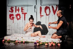 RISING. A woman in a slip sits on a mattress on the floor, while a man at the foot opens a can of drink. Behind the mattress is a panel with the words SHE GOT LOVE daubed on in red paint.