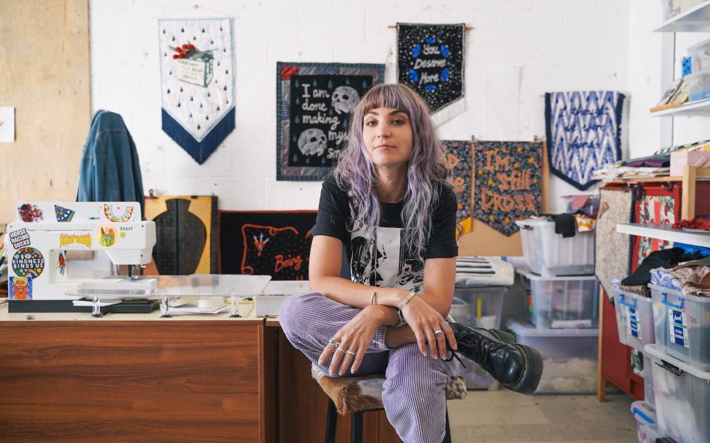 Textile banners by Tal Fitzpatrick will feature in the exhibition at MoAD. Photo: Supplied. A woman with wavy lavender coloured hair and a fringe sitting with her leg crossed on a stool inside a studio where her textiles works are displayed on the walls.