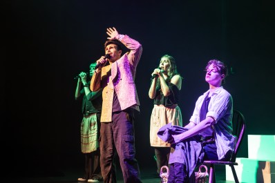 GRIT. Image is four young people on a stage against a black backdrop. A man at the front has a mic and is shielding his eyes from the light with his hand. Behind him two woman are also singing into mics, while a third woman is closer to the front sitting on a seat with crossed legs and lacing her fingers around her knee.