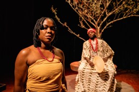 Akaraka. A dark set with two people of African appearance, a woman with braided hair and a wraparound shoulderless costume, wearing a red necklace and an older seated man, dressed in regal robes with a red hat.