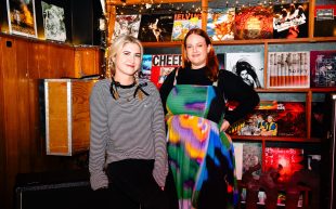 Casey O’Shaughnessy and Katie Rynne join BIGSOUND 2024 as Music Programmers. Photo: Jess Gleeson. Two Caucasian women standing inside a sound studio with vinyl covering a shelf to the right. Standing on the left is Casey with blonde plaits, a striped long-sleeve top and black paints. On the right is Katie with a coloured, heat-map like dress paired with black long sleeve underneath, she has amber hair.