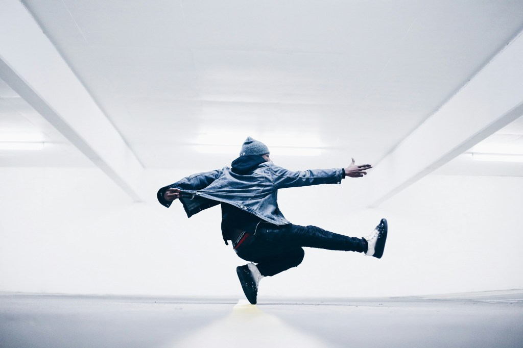 A dancer dressed in greay and black streetwear caught mid-move, balanced on the tip one foot with their other legs and both arms extended for balance. The background appears to an underground car park or similar liminal space.