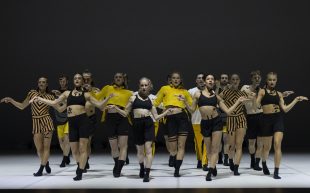 ‘Forever & Ever’ is a part of the triple bill ‘Ascent’ being toured by Sydney Dance Company this year. Photo: Supplied. An ensemble of 13 dancers on stage wearing black and yellow crop tops and shorts with black socks. The are in a walking stance with arms outstretched but slightly bent, looking powerful.