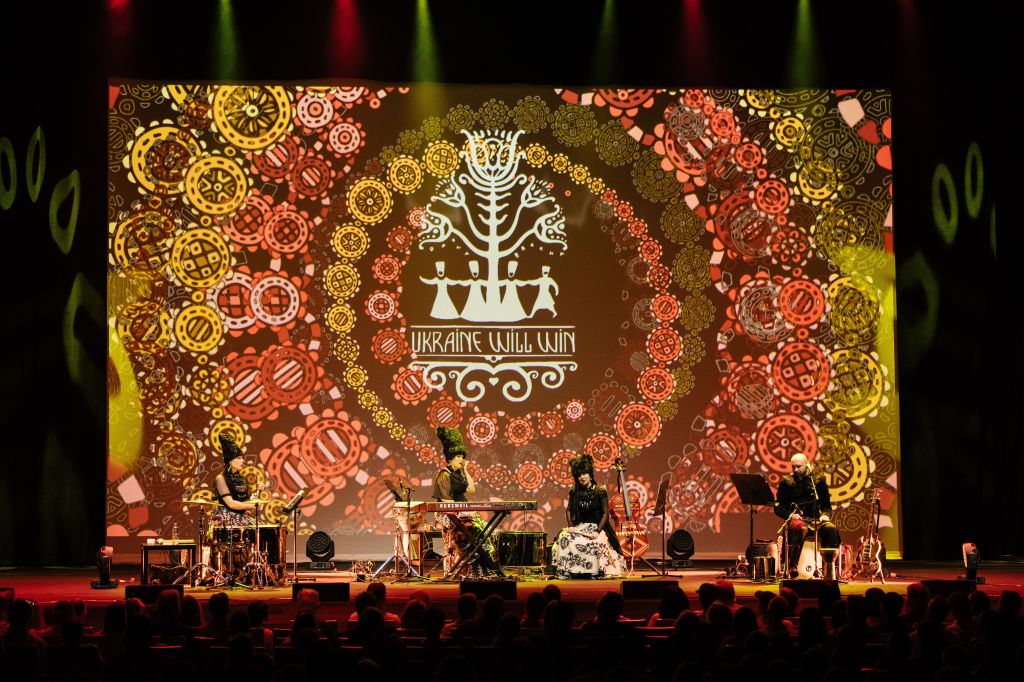 DakhaBrakha. Image is a concert stage with a huge circular abstract orange/yellow pattern on the back wall, and in front four black clad musicians, three of them women with tall black stovepipe hats and the fourth a man with a bald head.