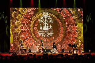 DakhaBrakha. Image is a concert stage with a huge circular abstract orange/yellow pattern on the back wall, and in front four black clad musicians, three of them women with tall black stovepipe hats and the fourth a man with a bald head.