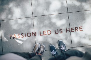 Two pairs of feet standing next to a sign on ground saying 'passion led us here'