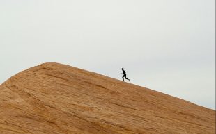 A runner in silhouette against the sky as they run up the steep slope of a large sand dune.