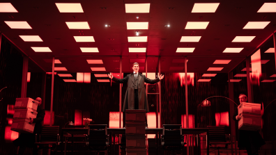 Lehmann Trilogy. Image is a man in a red tinted corporate office, with a grid of square fluorescent lights overhead. He is standing on a boardroom table with his arms outstretched to the side. Around him are file boxes and one is being carried out by an office worker.