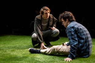 Milked. Two young men sit on a stage of fake grass, one looking down despondent, the other crouched in front of him.