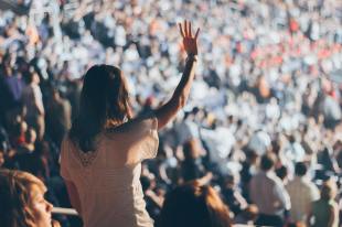 Consent. Image is a large audience with a person in the foreground of the shot with their back to the camera, standing and raising their hand.