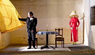 Private View. Image is of a man with Down's syndrome standing in a room looking out of a window and holding a billowing yellow curtain. In the middle of the room is a smalll round table with a lit candle on it and two chairs. In the right hand top corner is a female figure in a slinky figure hugging red dress, with a lampshade obscuring her face and head.