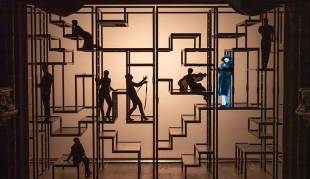 A scaffolding like set has several bodies positioned within it silhouetted against a lit backdrop. The Threepenny Opera.