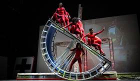Time Machine. STREB EXTREME ACTION. A group of red lycra clad performers cling to a huge half circle metal wheel in a display of dexterity and strength.