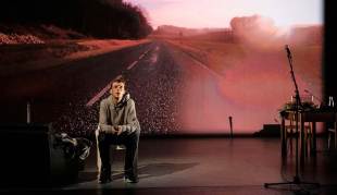 Qui a tué mon père. A man sits on a stage in front of a backdrop projection of an open road.