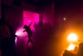 A person dances between pink and orange lights. The audience sits and stand around her to watch.