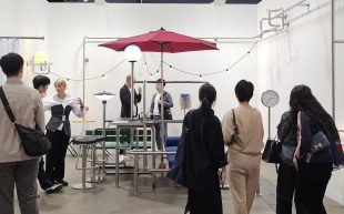 'In Transit' by Yona Lee at Fine Arts Sydney, Art Basel Hong Kong 2024, Discoveries section. Photo: ArtsHub. People gathering around a booth with stainless steel installations that make up a seemingly domestic setting, with benches, an outdoor umbrella, and fairy lights.