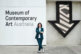 Woman in blue suit outside Museum of Contemporary Art Australia. To the right of her is a huge patterned arrow on the wall pointing down to the ground.