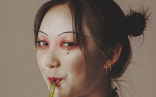 Whitesnake3000. A woman of Asian appearance looks at the camera from side on. She is smiling and wearing white face make-up while sucking on a green tube.