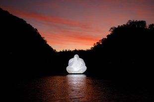A luminous inflatable statue of a contemplative male figures floats on a river at sunset. The sunset is reflected in the water and dark trees line the banks.