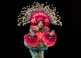A woman in bright, rainbow-colour floral crochet clothing with a headpiece, stands against a black background with her hands out.