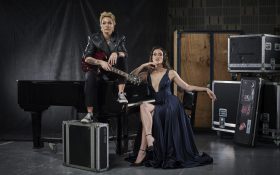 Australian Women in Music Awards 2024 open for nominations. Image: Sarah McLeod and Nina Korbe to perform at AWMA 2024. Photo: Guy Davies. McLeod has short cropped hair with smoky makeup, wearing a leather jacket, holding a bass and sitting on top of a piano. Beside her, Korbe is sitting, wearing a low neckline black dress with black heels. She has brown curly hair and a powerful expression. Both of them are presented in a powerful and beautiful manner against a backstage environment.