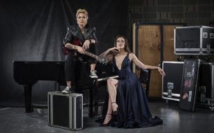 Australian Women in Music Awards 2024 open for nominations. Image: Sarah McLeod and Nina Korbe to perform at AWMA 2024. Photo: Guy Davies. McLeod has short cropped hair with smoky makeup, wearing a leather jacket, holding a bass and sitting on top of a piano. Beside her, Korbe is sitting, wearing a low neckline black dress with black heels. She has brown curly hair and a powerful expression. Both of them are presented in a powerful and beautiful manner against a backstage environment.