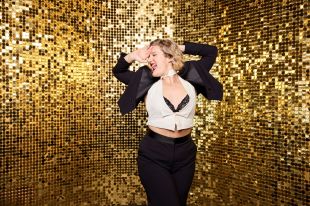 A white woman with short blonde hair poses dramatically before a gold lame screen. Her hands are behind her head, her face is turned to the right, and she is singing or screaming with joy. She wears a white shirt under a black suit, and her midriff and a hint of her black bra are visible.