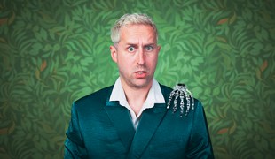 Against a green leafy wallpaper backdrop a white man with blond hair and a green jacket grimaces at the camera and has a spooky skeleton hand on his shoulder.