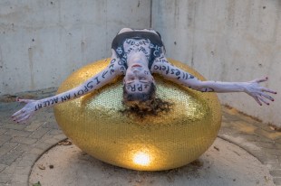 The PR.omised Land. Exhibition photo shows a blue painted woman with words written across her body lying on a golden egg-like sculpture.