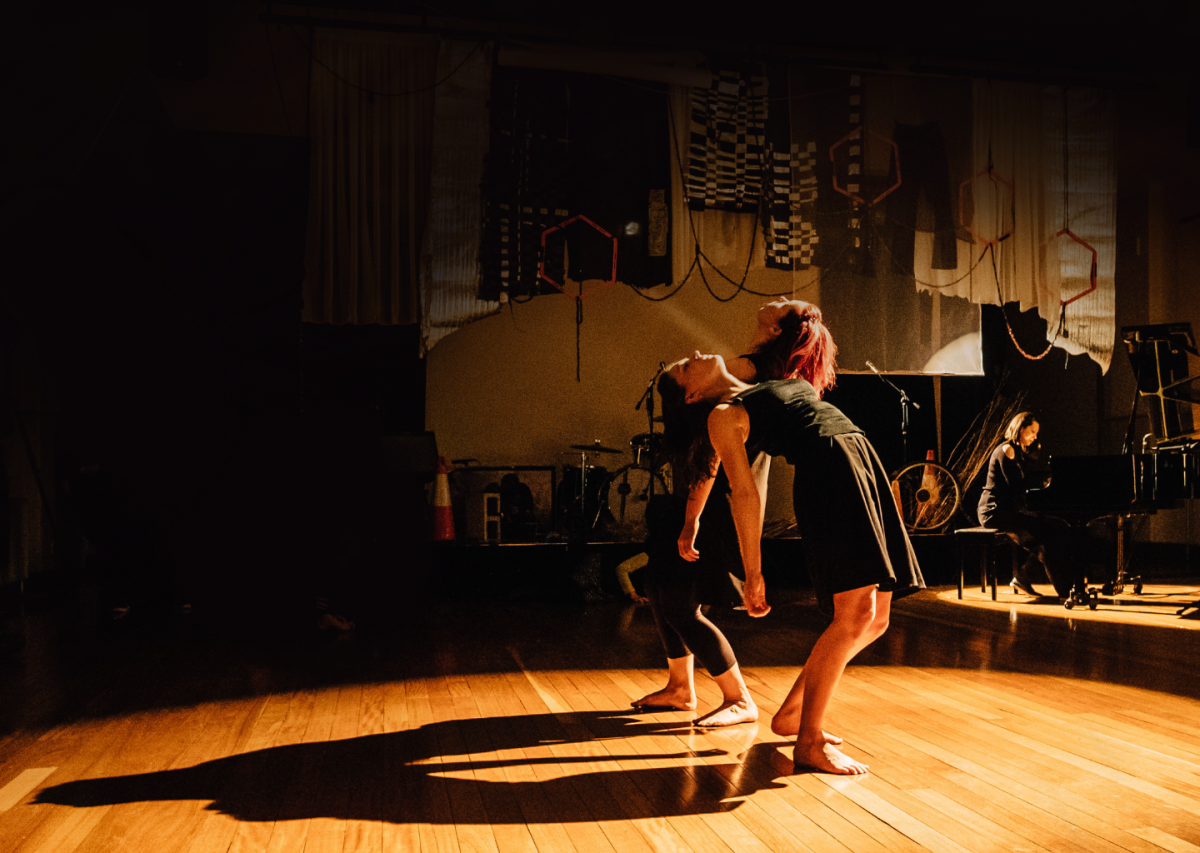 Two dancers lean back and look to the roof of the lowly lit studio. A pianist is in the background on the right.