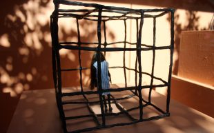 Jessica Nothdurft, 'The Cage', 2023, bronze, cotton and human hair. Installation view in 'Silly Girl' at artisan. Photo: ArtsHub. A small bronze figure with a blue striped dress, no facial expressions and long hair trapped inside a small bronze square cage.