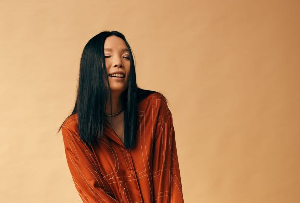 A woman of Asian appearance sits on a stool smiling at the camera. She has long dark hair and wears an orange-brown outfit. The background is indistinct.