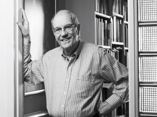 A black and white photograph of a well-presented, older white man standing in a doorway, with his left hand on his hip and hie right hand and upper arm braced against the doorframe. Many bookshelves are visible in the background behind them. Frank Van Straten.