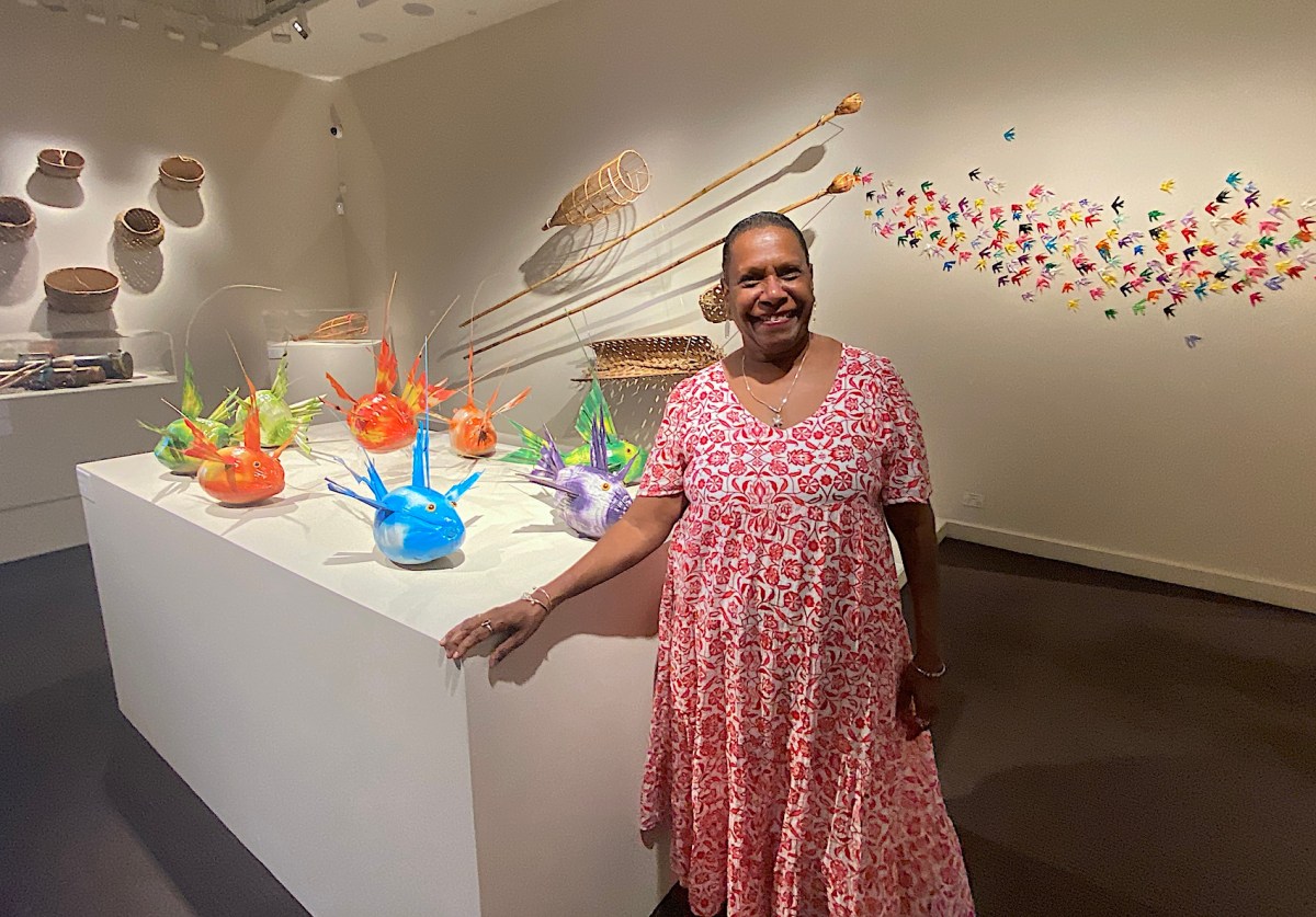 First Nations woman in red dress standing in gallery space with Torres Strait Island art. Gail Mabo.