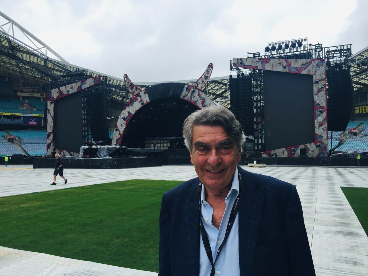 A grey-haired man wearing a blue shirt and a dark blue suit, as well as a lanyard around his neck, smiles at the camera. He is standing on the pitch of a sports stadium and a large soundstage has been erected behind him.