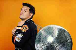 A side on picture of a man in a black jacket with colourful patches on the sleeve, lugging a large mirror ball over his shoulder.
