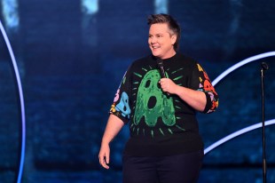 Hannah Gadsby. Image is a woman with short hair and a black jumper with a big green image of an open mouthed ghost on the front. She is holding a microphone and smiling.