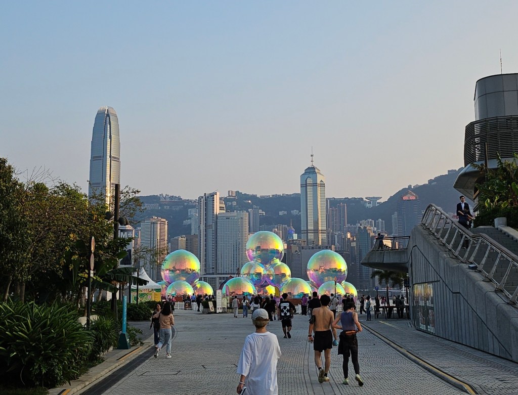 ‘Ephemeral’ by Sydney-based design studio Atelier Sisu at WestK FunFest. Photo: ArtsHub. Art precincts. Large iridescent installations that resemble bubbles are placed in three clusters outdoors with the background of the cityscape of Hong Kong island, featuring tall buildings and water.