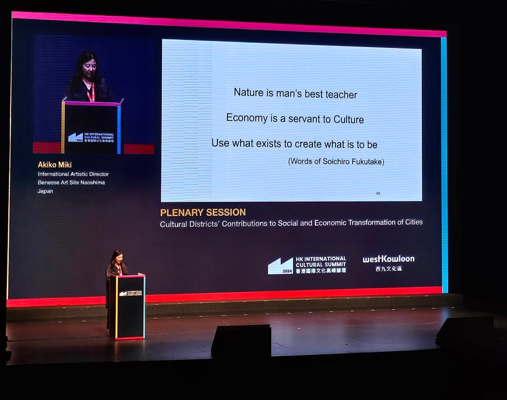 Akiko Miko speaking at Hong Kong International Cultural Summit 2024. Photo: ArtsHub. Arts Precincts. Miko is standing in front of a large projected slide with the sentences ‘Nature is man’s best teacher / Economy is a servant to Culture / Use what exists to create what is to be / (Words of Soichiro Fukutake)