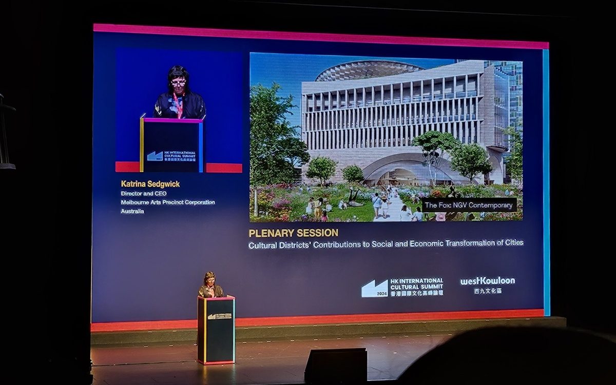 Katrina Sedgwick is standing on stage in front of a large projector screen presenting a slide that includes an architectural render of The Fox: NGV Contemporary.